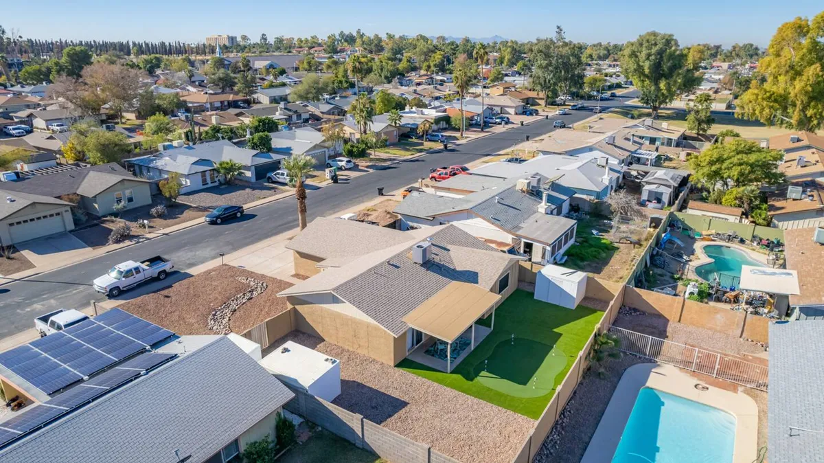 This image shows a vibrant backyard scene featuring a sparkling pool, lush green lawn, and modern homes in the background. The space is clearly designed for social gatherings, relaxation, and Instagram-worthy moments in the sun. Solar panels on the roof hint at modern amenities.