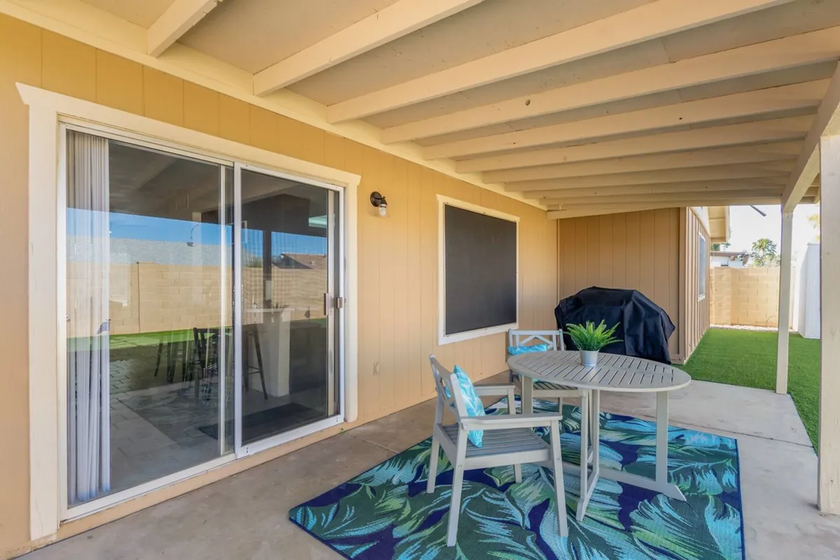 A casual and welcoming patio scene featuring a dining table with chairs, a potted plant, and a grill cover set against a backdrop suitable for sunset dinners or morning coffee. The ambiance is ideal for outdoor dining and relaxing with friends.