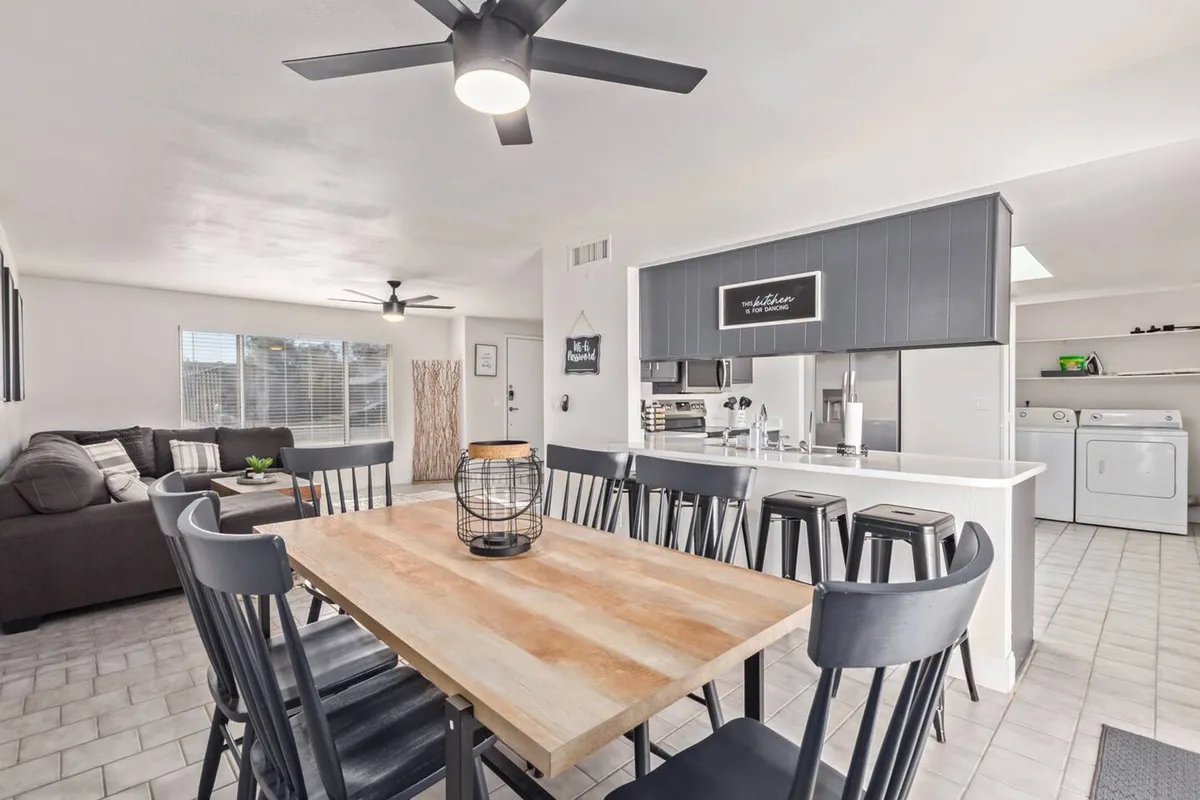 An airy interior shot encompassing a dining table, sofa, open kitchen area with modern appliances, and a ceiling fan. The space feels interconnected and lively—ideal for group gatherings, meals, and casual conversations.