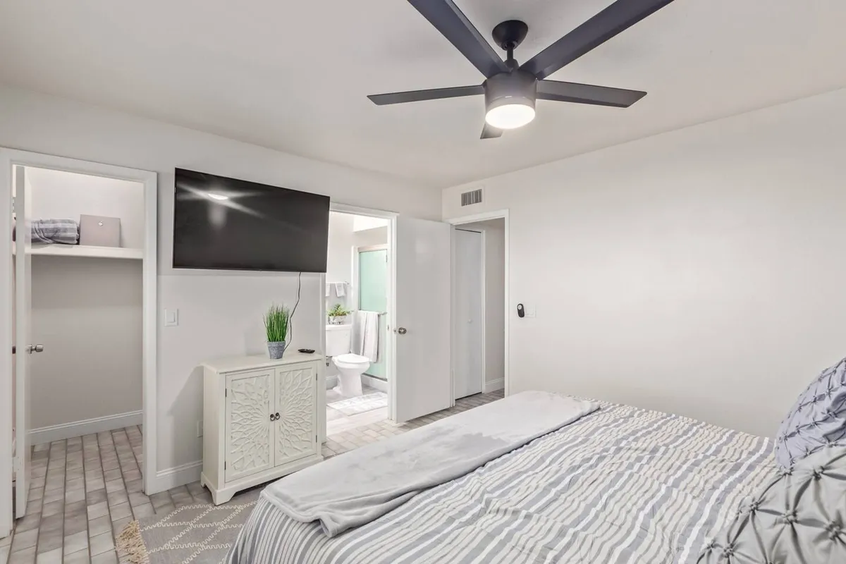 A contemporary bedroom with a comfortable bed, ceiling fan, and a large television prominently placed for easy viewing. The scene also includes a closet and direct access to a bathroom, reinforcing the idea of convenience and relaxation.