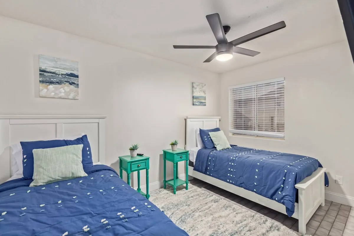 A cozy interior room with two twin beds, green accent tables, ceiling fan, and playful wall art. The space is relaxed and inviting, perfect for children or siblings to share a restful night's sleep after a day of adventure.