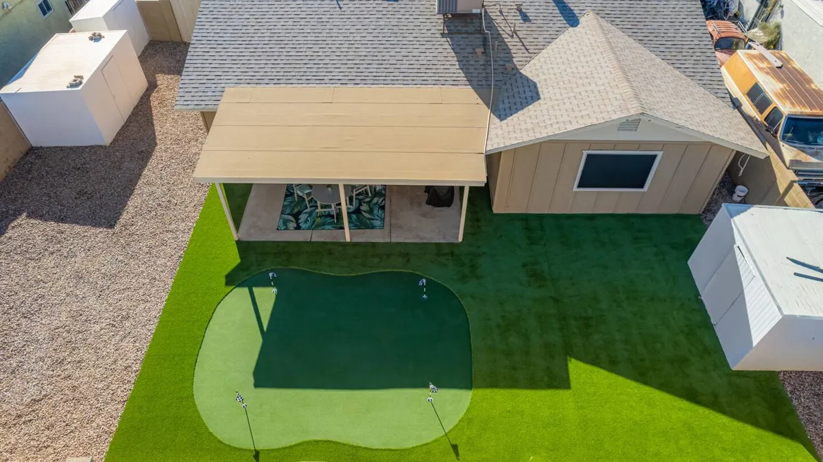An inviting exterior view highlighting an artificial putting green, a covered patio area, and a lush green lawn. The space is recreational and ideal for both golf practice and family gatherings, with shade for relaxing and plenty of room for activities.