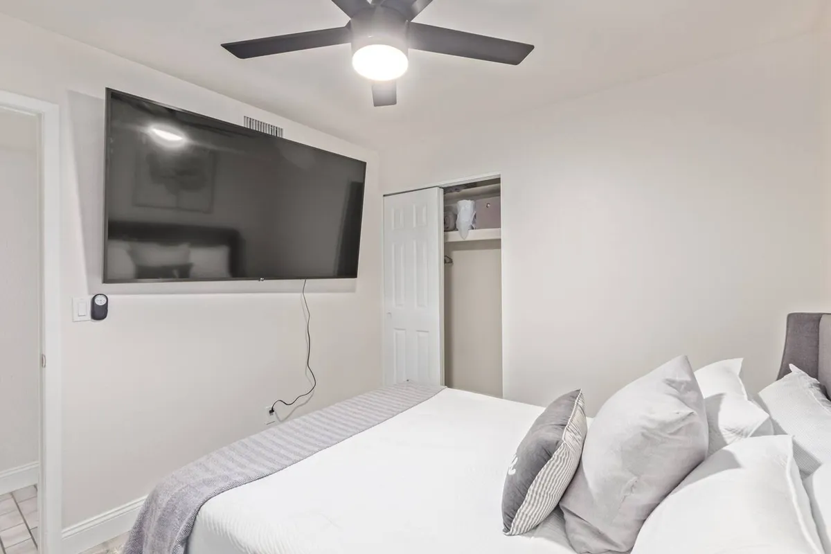 A cozy bedroom with a large flat-screen TV mounted opposite a neatly made bed adorned with multiple pillows. The space is decorated in soothing tones and features a ceiling fan, giving it a welcoming and relaxing ambiance ideal for enjoying entertainment from the comfort of bed.