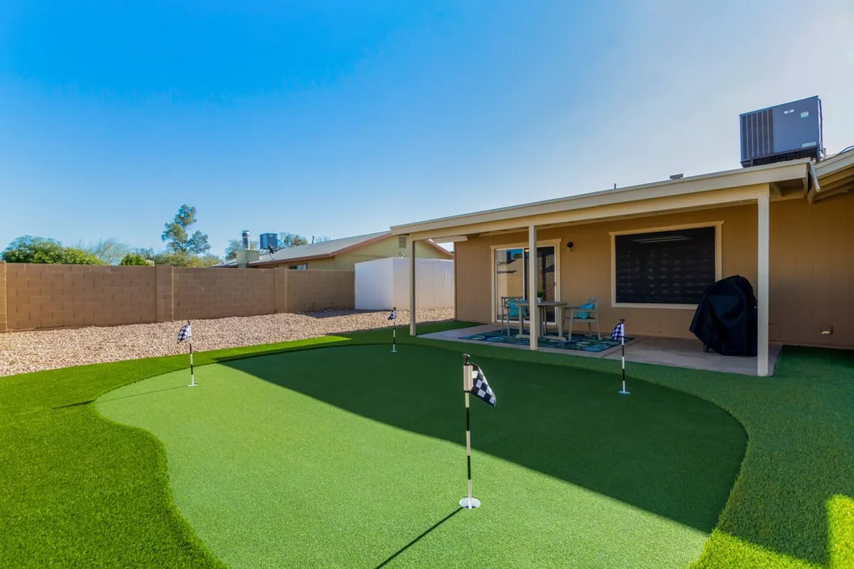 The image highlights a backyard with a putting green, cozy outdoor seating, and an inviting house facade. The scene suggests sunny afternoons spent practicing golf with friends, or relaxing outdoors in a social setting.