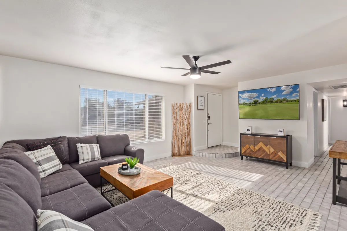 This image features a spacious interior with a large sectional sofa, a coffee table, a sleek television mounted on the wall, and a decorative plant, all bathed in cozy natural light. The arrangement provides an inviting atmosphere ideal for relaxing or entertaining guests.