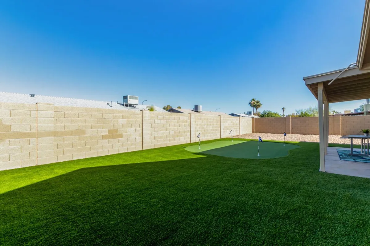 A sunny backyard scene featuring a manicured lawn, a putting green, and a spacious patio area. The mood is cheerful, with open sky and plenty of space for kids to run around or families to gather for outdoor fun.